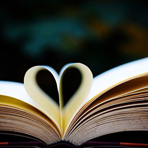 Aufgeschlagenes Buch, zwei Seiten zu einem Herz geformt, vor unscharfem dunklem Hintergrund.